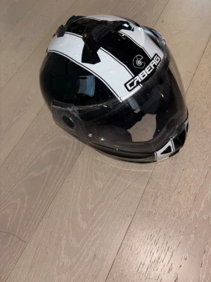 Svartvit motorcykelhjälm Caberg - Snygg svart och vit motorcykelhjälm från Caberg med visir och modern design. Hjälmen har ventilationsöppningar och tydlig logga framtill. Perfekt för dig som vill ha både stil och funktion på vägen.