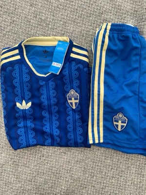 Sverige fotbollskit Adidas blå/gul för 10 åring - Säljer ett blått och gult Sverige fotbollskit från Adidas med retroinspirerat mönster. Tröjan har V-ringning, gula detaljer och broderad Sverige-logga samt Adidas-märke. Matchande shorts med gula ränder och Sverige-emblem. Perfekt för fotbollsträning eller match.