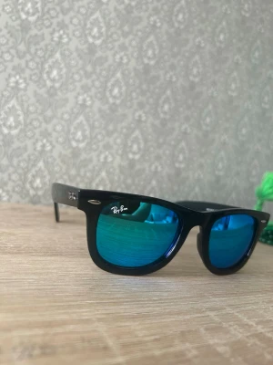 Ray-Ban svarta solglasögon blå lins - Snygga svarta solglasögon från Ray-Ban med klassisk wayfarer-form och spegelblå linser. Ikonisk logga på ena linsen och skalmen. Perfekta för dig som vill ha en cool och tidlös look i solen.