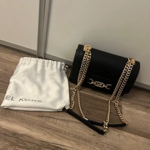 Svart axelväska från Michael Kors - Svart axelväska från Michael Kors i slät läderlook med guldfärgade kedjor och MK-detalj framtill. Väskan har en stilren, rundad form och levereras med en vit dustbag. Väskan har ett fack på baksidan och två inuti. Säljes då den tyvärr aldrig kommit till användning. 