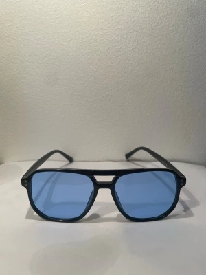 Blåa solglasögon  - Snygga solglasögon med svart båge och rektangulära blåtonade glas. Klassisk design som ger en cool vibe och passar perfekt till sommaren. Lätta att matcha med olika outfits och ger ett trendigt intryck.