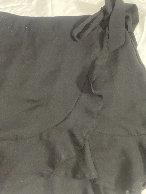 Svart omlottkjol med volang - Snygg svart kort omlottkjol med volangkant och knyt i midjan. 