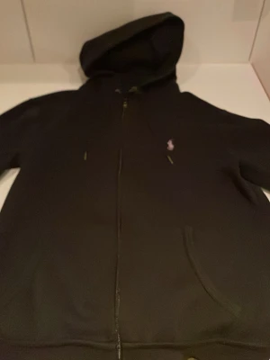Svart hoodie från Polo Ralph Lauren - Svart hoodie med dragkedja från Polo Ralph Lauren. Klassisk design med huva, fickor framtill och den ikoniska broderade loggan på bröstet. Perfekt för en avslappnad och stilren look. Mjuk och skön bomullsblandning.
