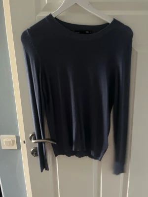Mörkblå stickad tröja - En stilren mörkblå stickad tröja med rund halsringning och långa ärmar. Tröjan har en klassisk passform och ribbstickade muddar vid ärmslut och nederkant. Perfekt att matcha med jeans eller kjol för en enkel och snygg look.
