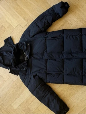 Svart pufferjacka från DIYUFEI XL - Säljer en svart pufferjacka från DIYUFEI i storlek XL. Jackan har en hög krage, avtagbar huva och stora quiltade partier. Dragkedja och knappar framtill samt ett coolt mönstrat innerfoder. Perfekt för kalla vinterdagar.