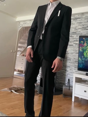 Klassisk svart kostym herr strl 50 - Säljer en stilren svart kostym med kavaj och matchande byxor. Kavajen har två knappar och klassisk passform, perfekt till skjorta och slips. Materialet känns mjukt och följsamt, troligen polyester eller ullblandning. Enkel och tidlös design utan mönster. Använd endast 1 gång vid studenten! 