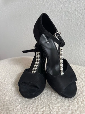Svarta pumps med pärldetaljer som nya - Snygga svarta pumps från Graceland med öppen tå och T-rem dekorerad med vita pärlor. Skorna har en justerbar ankelrem med spänne och en klassisk klack. Materialet ser ut att vara mockaimitation som ger en elegant look. Använd en gång.