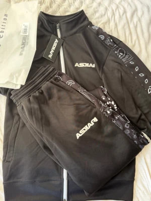Svart tracksuit från Askari - Säljer en svart tracksuit från Askari med vita logotyper på både tröja och byxor. Setet har bandanamönstrade detaljer längs ärmar och ben, dragkedja framtill och elastisk midja på byxorna. Perfekt för streetwear vibes eller tränaingskläder. Den är helt ny jag testade den på mig en gång. Passar både s och m. Den finns inte längre (limited edition). 