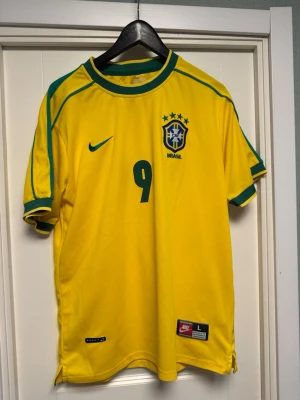 Brasilien Ronaldo 9 matchtröja Nike - Gul Brasilien fotbollströja från Nike med gröna detaljer, nummer 9 och Ronaldo på ryggen. Klassisk landslagströja med broderat landslagsmärke och Nike-logga. Perfekt för dig som älskar fotboll och vill hylla en ikon.