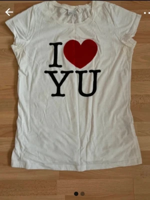 Vit t-shirt med I Love YU-tryck - Vit t-shirt med korta ärmar och tryck framtill med texten 'I ♥ YU' i svart och rött. Klassisk passform och rund halsringning. Perfekt för en avslappnad och personlig stil. Förminskad somM/L