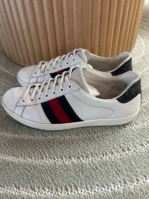 Gucci ace - Snygga vita sneakers från Gucci med klassiska röd och blå ränder på sidan och svart detalj vid hälen. Skorna har låga skaft, vita snören och är tillverkade i läder. Perfekta för dig som gillar stilrena och ikoniska märken.