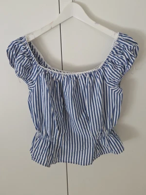 Blåvit randig singoallablus med puffärm - Söt blus med blå och vita ränder, puffiga korta ärmar och resår i midjan. Urringad hals med spetsdetalj. Perfekt för en somrig look och lätt att matcha med jeans eller kjol.