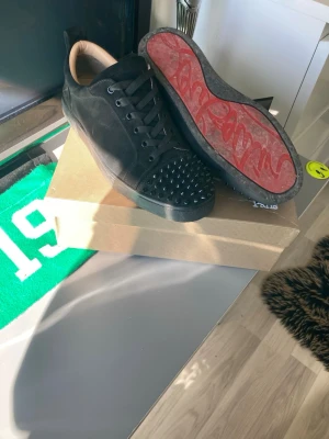 Svarta Christian Louboutin sneakers - Säljer ett par svarta sneakers från Christian Louboutin med ikoniska röda sulor och nitar på tån. Skorna har lågt skaft, svarta skosnören och är tillverkade i mocka och läder. Perfekta för dig som vill sticka ut med exklusiv design och edgy detaljer. Låda och dustbag ingår.