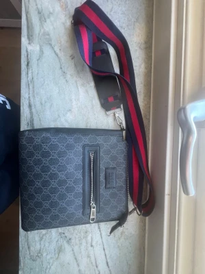 Gucci axelväska med monogram - Snygg axelväska från Gucci i mörkgrått med klassiskt GG-monogram över hela väskan. Väskan har en framficka med dragkedja och en bred axelrem i rött och marinblått. Materialet är canvas med detaljer i läder. Perfekt för dig som gillar streetstyle och designer.