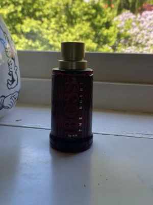BOSS The Scent Elixir parfym - En elegant parfym från Hugo Boss, The Scent Elixir. Flaskan är mörkröd med guldigt lock och har en stilren, cylindrisk form. Texten 'BOSS' och 'THE SCENT' syns tydligt på flaskan. Perfekt för dig som gillar exklusiva dofter och snygg design. Liter mer en hälften kvar!! Ingen box!