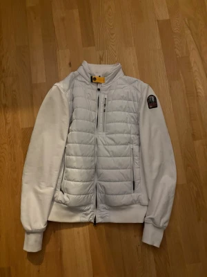 Vit pufferjacka från Parajumpers - Snygg vit pufferjacka från Parajumpers med quiltad front och rygg, samt släta ärmar. Jackan har hög krage med gul detalj, flera dragkedjefickor och en patch på ena ärmen. Perfekt för kalla vinterdagar.