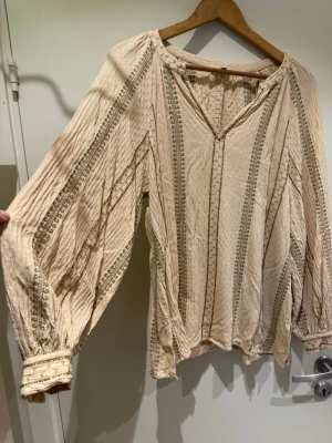 Super fin! Beige boho-blus från Indiska - Så fin! Luftig beige blus från Indiska med boho-inspirerade broderier och pärlbrodyr. Blusen har v-ringning och långa lite puff ärmar med knappar vid handlederna. Perfekt för en avslappnad och trendig look. Skönt material i viskos. Stl M, ganska liten i stl för att vara indiska. Använd någon enstaka gång och tvättad 1 ggr så i mycket fint skick. Möjligen lite skrynklig. Nypris 999:-