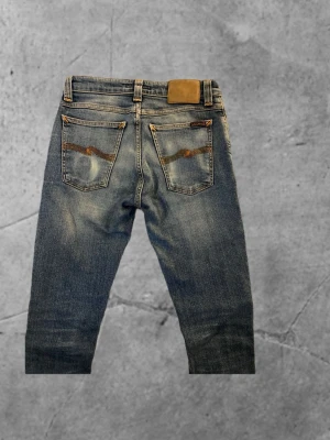 Blå jeans från Nudie Jeans - Säljer ett par blå jeans från Nudie Jeans med klassisk femficksdesign och orangea sömmar. Jeansen har låg midja och är tillverkade i stretchig bomull. Snygga slitningar och Nudie Jeans logga på bakfickan ger en cool vibe. Skriv vid minsta lilla fråga!🙋‍♂️ 