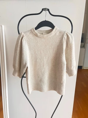 Beige stickad tröja med puffärm - Snygg beige stickad tröja med korta puffärmar och ribbade muddar. Tröjan har en rund halsringning och är gjord i ett mjukt material som känns skönt mot huden. Perfekt för dig som gillar en stilren och trendig look.