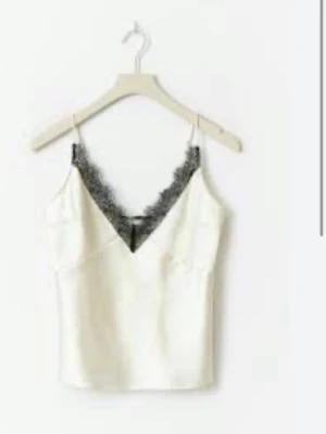 Vit linnetopp med spets från Gina Tricot - Supersnygg vit linnetopp från Gina Tricot med tunna axelband och svart spetsdetalj vid v-ringningen. Perfekt att styla med jeans eller kjol för en trendig look. Topp i satinliknande material som känns lyxigt och mjukt.