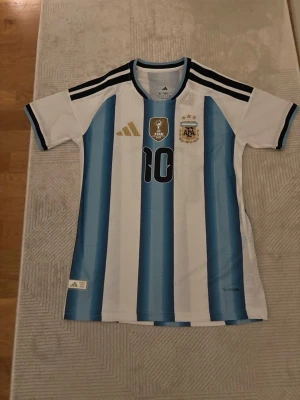 Argentina Messi matchtröja Adidas - Säljer en officiell Argentina fotbollströja från Adidas med Messis namn och nummer 10. Tröjan är vit med ljusblå ränder, svarta detaljer och guldiga loggor på bröstet. Korta ärmar och klassisk sportig passform. Perfekt för fotbollsfans!