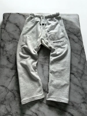Gråa Essentials mjukisbyxor - Snygga grå mjukisbyxor från Fear of God Essentials med svart logga på vänster lår. Byxorna har resår i midjan med dragsko och raka ben. Perfekta för en chill och avslappnad stil. Tillverkade i mjukt material för maximal komfort. Uncuffed, skick 8/10 finns märke på snöret som såkert går bort i tvätten.