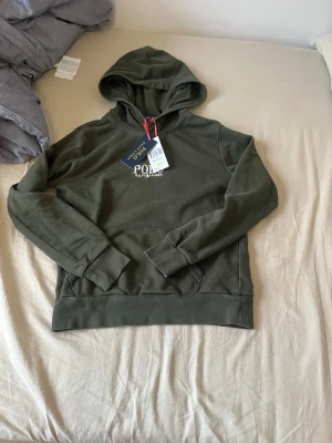 Olivgrön hoodie från Polo Ralph Lauren - Olivgrön hoodie från Polo Ralph Lauren med klassisk huva och magficka. Tröjan har ribbade muddar vid ärmslut och nederkant samt diskret logga på bröstet. Tillverkad i mjuk bomullsblandning för en skön känsla. Ny pris 1300 