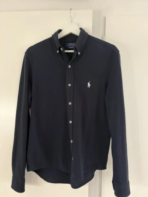 Mörkblå skjorta från Polo Ralph Lauren  - Klassisk mörkblå skjorta från Polo Ralph Lauren med button-down krage och vit broderad logga på bröstet. Skjortan har långa ärmar, knappar framtill och är tillverkad i featherweight mesh. Perfekt för en stilren look.