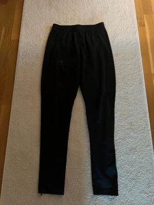 Svarta träningsbyxor från Under Armour - Svarta träningsbyxor från Under Armour med smal passform och resår i midjan. Byxorna har dragkedja vid bensluten och är tillverkade i ett lätt och stretchigt material som passar perfekt för sport och träning.