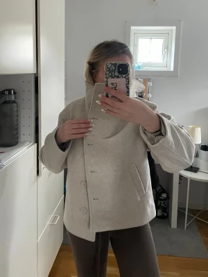 Beige kappa med hög krage - Stilren beige jacka ”Blaire” fråm Lager 157 med hög krage och knäppning framtill. Jackan har en rak, loose passform. Perfekt för dig som gillar minimalistisk och modern stil. Den är helt oanvänd , i storlek XS. Säljer för att det tyvärr inte satt bra på mig. 