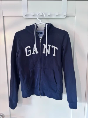 Marinblå hoodie från GANT Storlek S - Snygg marinblå hoodie från GANT med dragkedja framtill och vita snören i huvan. Stor GANT-logga i vitt på bröstet och två fickor fram. Tillverkad i mjukt bomullsmaterial.