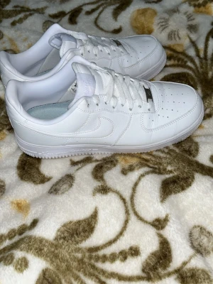 Nike Air Force 1 vita sneakers - Klassiska helvita Nike Air Force 1 sneakers i lågt snitt. Skorna är tillverkade i läder med perforerad tå för ventilation och har Nike-logga på sidan samt AIR-detalj på hälen. Snygg och tidlös modell som passar till det mesta.