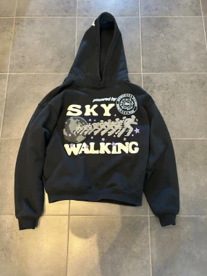 Svart hoodie Broken Planet Sky Walking - Snygg svart hoodie från Broken Planet med stor huva och trycket 'SKY WALKING' i vitt och lila på framsidan. Tillverkad i 100% ekologisk bomull. Hoodien har ribbade muddar och coolt grafiskt motiv med stjärnor och figurer.