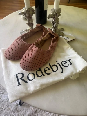 Rosa perforerade ballerinaskor Rodebjer - Snygga ballerinaskor från Rodebjer i en mjuk, dammigt rosa nyans. Skorna är tillverkade i läder med perforerat mönster och har elastisk kant för skön passform. Perfekta för dig som gillar stilrena och trendiga detaljer. Slutsålda och dyra i inköp, närmare 3000:- sista bilden visar hur de derbyt på knutna (dock i annan färg) 