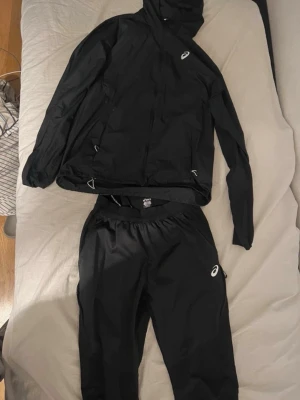 Svart Jacka från Asics - Säljer ett svart jacka från Asics. Jackan har huva, dragkedja och justerbara snören i nederkant. Perfekt för löpning eller annan träning utomhus.