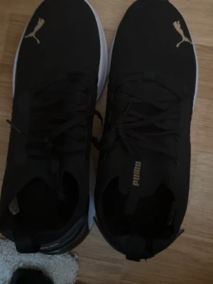 Svarta Puma sneakers med vit sula - Snygga svarta sneakers från Puma med vit, tjock sula och guldfärgade Puma-loggor på tån och plösen. Skorna har snörning och är tillverkade i textil och syntetmaterial. Perfekta för en sportig och trendig look. Har använt skorna ett par gånger i gymmet, därför ser sulorna ut så. Men inget som påverkar funktionen. 