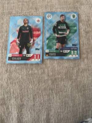 Topps fotbollssamlarbilder - Två fotbollssamlarbilder från Topps med spelare i matchställ. Ena kortet visar en målvakt från Red Bull Salzburg och det andra en utespelare från Sporting CP. Korten har färgstarka bakgrunder och tydliga klubb- och spelarnamn. Perfekt för samlare av fotbollskort.