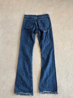 Blå bootcut jeans med broderi - Unika blå bootcut jeans med råa kanter och broderade blommor längs ena benet. Jeansen har klassisk femficksmodell och är tillverkade i denim. Perfekta för dig som vill sticka ut med en kreativ och personlig stil.