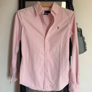 Rosa randig skjorta från Ralph Lauren - Köptes för 550kr. Klassisk rosa och vit randig skjorta från Ralph Lauren med broderad logga på bröstet. Skjortan har lång ärm, knappar framtill och är i modellen Custom Fit. Perfekt för en stilren och tidlös look.