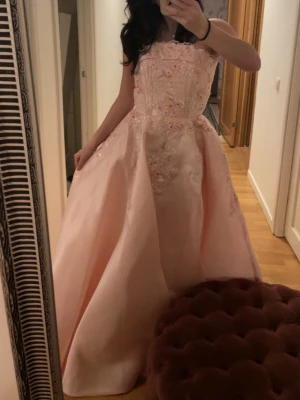 Ljusrosa balklänning med blommor - Superfin ljusrosa långklänning med axelbandslös design och utsvängd kjol. Klänningen har vackra blommiga broderier och pärldetaljer upptill, perfekt för bal. Materialet känns lyxigt och ger en prinsesskänsla. Storlek xs men har corset som kan anpassa storleken 