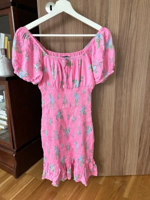 Rosa blommig kortklänning med volang - Söt rosa kortklänning med blommigt mönster och volang nertill. Klänningen har puffiga korta ärmar och smockad midja för en figurnära passform. Perfekt för sommarens alla tillfällen.