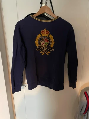 Marinblå tröja från Polo Ralph Lauren - Snygg marinblå långärmad tröja från Polo Ralph Lauren med broderat emblem i guld, rött och grönt på ryggen. Tröjan har ribbade muddar och krage med gula detaljer. Perfekt för en preppy stil.