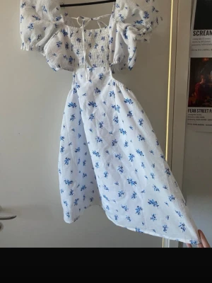 Vit klänning med blå blommor H&M - Söt vit klänning från H&M med blått blommönster, puffiga korta ärmar och smockad överdel. Klänningen har cut-out detaljer i midjan och knytning fram. Perfekt för sommarens varma dagar och har en luftig, romantisk stil.