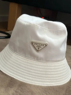 Vit bucket hat från Prada - Snygg vit bucket hat från Prada med klassisk logga framtill. Hatten är tillverkad i polyester med bomullsfoder, vilket gör den både lätt och bekväm. Perfekt accessoar för att lyfta din stil och ge en exklusiv touch till din outfit.