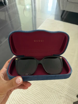 Gucci solglasögon med blått fodral - Säljer ett par stilrena solglasögon från Gucci. De kommer i ett lyxigt blått fodral med vinröd insida och guldfärgad Gucci-logga. Perfekt accessoar för dig som vill ha något exklusivt och trendigt.