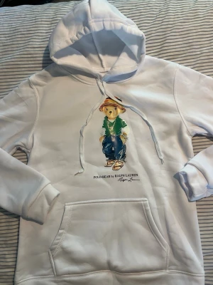 Vit hoodie Polo Bear Ralph Lauren - Vit hoodie från Ralph Lauren med ikoniska Polo Bear-trycket på bröstet. Hoodien har huva med dragsko och stor magficka framtill. Materialet är mjukt och bekvämt, perfekt för en avslappnad stil. Snygg detalj med färgglad björn i streetwear.