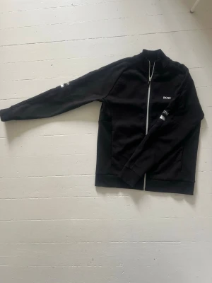 Svart zip-tröja från BOSS - Svart stilren ziptröja från Hugo Boss med silvriga logotyper på ärmar och bröstet. Storlek S - fint skick.