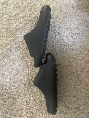 Svarta yeezy tofflor med sågtandssula - Säljer ett par svarta Adidas tofflor i skön och mjuk gummimaterial. De har en chunky sågtandad sula och stilren design med Adidas-logga på fotbädden. Perfekta för chill dagar hemma eller till stranden.