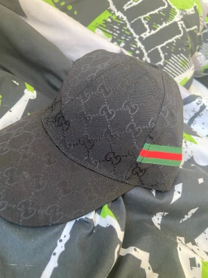 Svart keps - Snygg svart keps  med klassiskt över hela kepsen. På sidan finns en grön och röd rand som ger en ikonisk touch. Perfekt accessoar för att lyfta din streetwear-look.