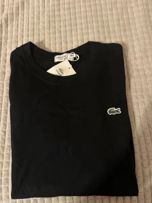 Svart t-shirt från Lacoste - Snygg svart t-shirt från Lacoste med klassisk rund halsringning och diskret logga på etiketten. Perfekt basplagg med enkel och stilren design som funkar till allt. Regular fit för en bekväm passform.